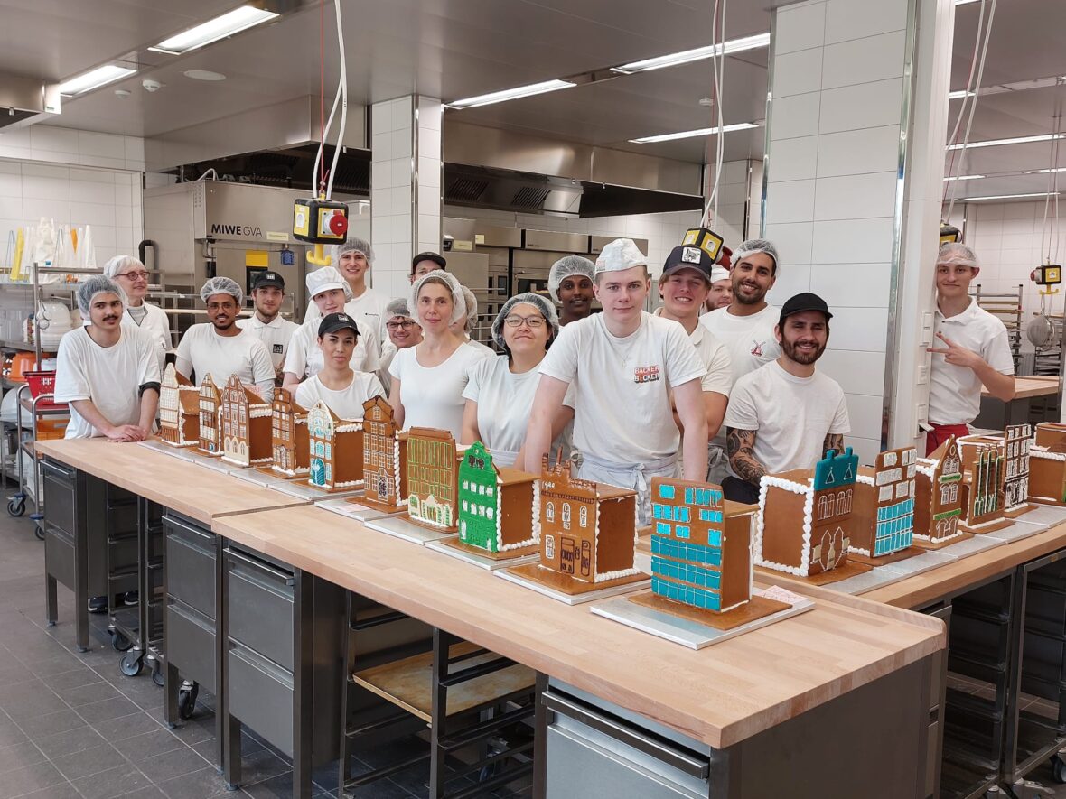 Junge weiß gekleidete Personen stehen hinter einem Tisch voller dekorierter Lebkuchenhäuser
